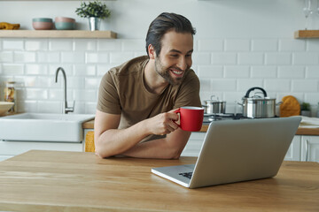 Happy young man using laptop and holding red cup while leaning at the kitchen island
