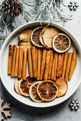 Obraz na płótnie Canvas Dried citrus slices and cinnamon sticks in white bowl with fir green and pine cones. Winter flavor from fruits and spices. Top view.