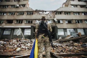 Deurstickers Oekraïense militaire vrouw met de Oekraïense vlag in haar handen op de achtergrond van een geëxplodeerd huis © alimyakubov