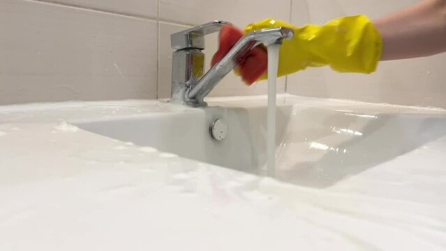 Close-up of a hand covered with a yellow rubber glove wiping a sink with a red sponge and running water
