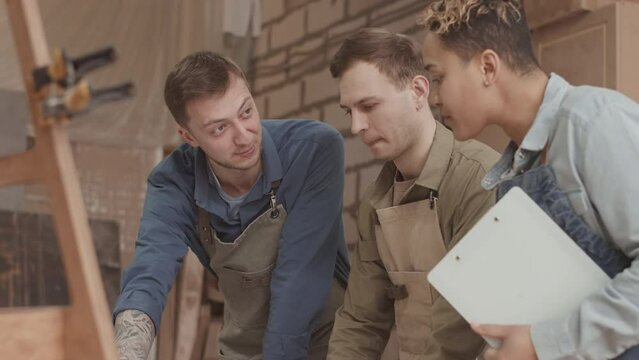 Team of three male and female carpenters in aprons looking at drawing plan on wooden desk while discussing construction in workshop