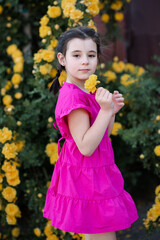 Stylish little kid girl 6-7 year old smelling yellow rose flower over blooming bushes in garden outdoor close up. Looking at camera. Springtime. Cute child toddler over nature background. Childhood.