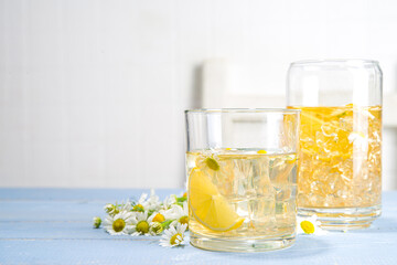 Herbal organic cold drink. Infused flower water. Iced daisy Chamomile tea or lemonade with lemon slice and ice cubes, in two different glasses, blue wooden background copy space