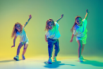Group of children, little girls in sportive casual style clothes dancing in choreography class isolated on green background in yellow neon light. Concept of music, fashion, art