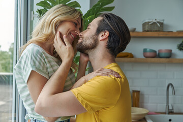 Beautiful young couple embracing other while standing near window at the kitchen