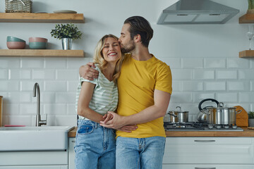Cute young couple embracing and kissing while standing at the domestic kitchen