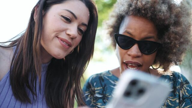 Two smiling multiethnic women talking and looking at phone while sitting on the bench outdoors