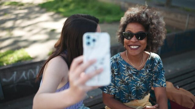 Two cheerful multiethnic women making selfie by phone while sitting on the bench outdoors