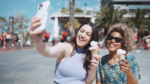 Two smiling multiethnic women making selfie with ice cream on phones camera outdoors