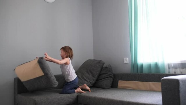 Funny toddler girl plays with pillows on couch, builds house and throws. Happy childhood, cozy interior
