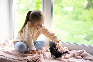 A cute girl plays with her little fluffy kitten sitting on the windowsill of the house. Friendship...