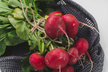 Bunch of fresh dirty organic ripe red radish with green leaves in eco-friendly reusable produce bag on white background. Healthy grocery, vegan raw food and ingredients for cooking concept. Close-up