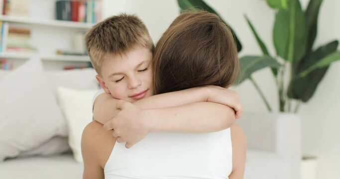 Young mother and her son have fun at home in the white living room. Close-up of boy hugging and kissing his mom. Active family, parent and child spend time together enjoying fun activities.