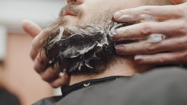 Male barber's hands wash beard of a Caucasian man with shampoo, close-up. Professional hands of the master massage the hipster's beard by washing it