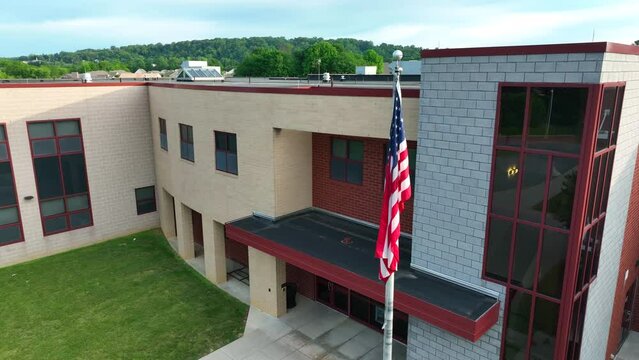 Aerial orbit around stagnant American flag. Flag hoisted on flagpole lays still in front of American elementary school.