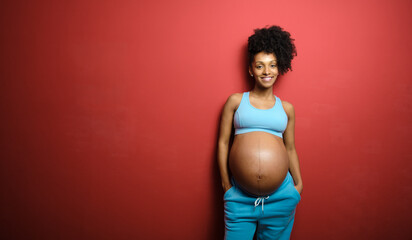 Portrait of blissful black pregnant woman on fitness sportswear against red background.
