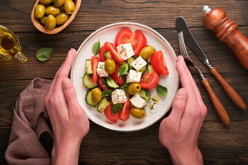 Greek salad. Fresh Greek salad with fresh vegetables, tomato, cucumber, green olives, feta cheese and man hands on old dark wooden table background. Top view.