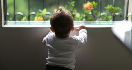 Cute baby peeking outdoors during rainy day. Infant toddler standing by window looking out - obrazy, fototapety, plakaty