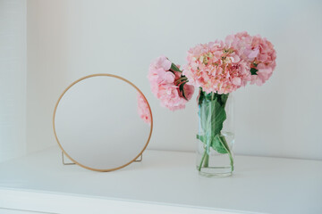 Cute small round cosmetic mirror in golden frame and glass vase with hydrangeas flowers on...