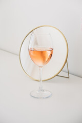 Cute small round cosmetic mirror in golden frame and glass of rosé wine on dresser..White interior. Decor details.
