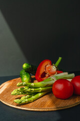 Vegetables lie on a wooden board: tomatoes, asparagus, cucumbers, red bell peppers. brown, dark gray background. place for text.