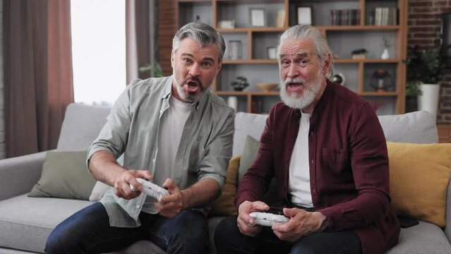 Adult son and senior father having fun, enjoy spending time together. Two generations family playing video games using gamepads at home. Fathers Day. Active modern elderly people.
