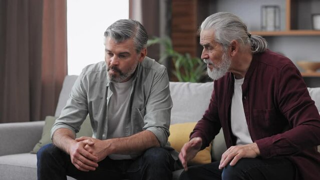 Family conflict. Adult son visiting senior father. Two generations family has unpleasant conversation sitting on couch at home. Generational confrontation.