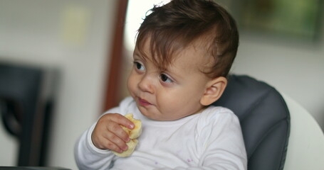 Cute baby boy eating pieces of banana seated on highchair