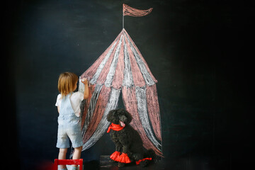 Kid with dog poodle draw circus tent on black chalk board, concept of children's creativity and imagination, copy space