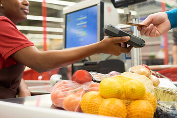 Young black woman holding payment terminal while buyer holding credit card while paying for bought food products in supermarket - obrazy, fototapety, plakaty