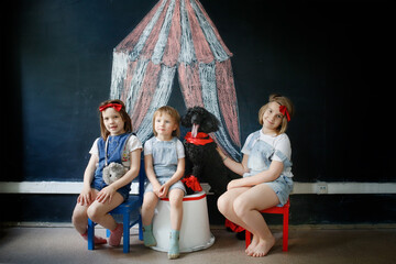 Children together with pet dog play in theater, circus. Black poodle and three European kids against background of black chalk board with circus pattern.
