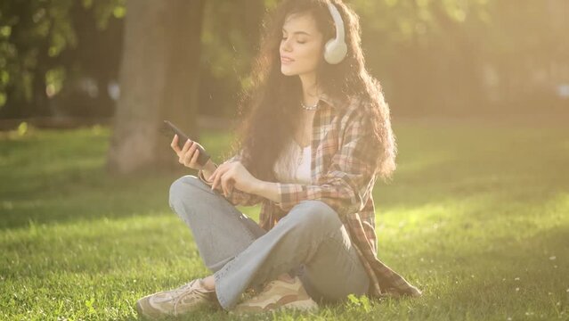 Photo of a cheerful optimistic beautiful young curly woman sitting on grass in park outdoors listening music with headphones using mobile phone
