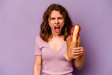 Young caucasian woman eating a sandwich isolated on purple background screaming very angry and...