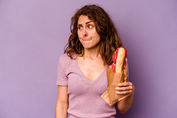 Young caucasian woman eating a sandwich isolated on purple background confused, feels doubtful and...