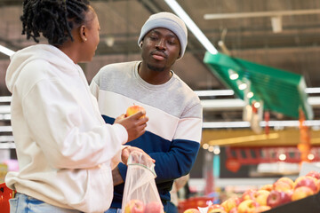 Fototapeta na wymiar Young man with shopping cart looking at fresh apple held by his girlfriend choosing fruits in grocery department of supermarket