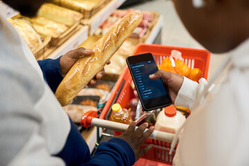 Shopping list on screen of smartphone held by young African American woman over cart with fresh food products from grocery department