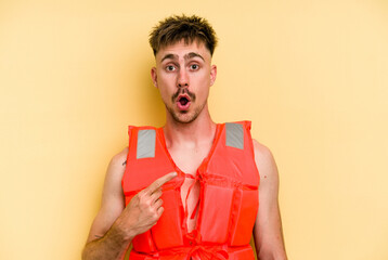 Young caucasian man wearing a life jacket isolated on yellow background pointing to the side