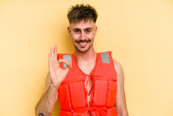 Young caucasian man wearing a life jacket isolated on yellow background cheerful and confident...
