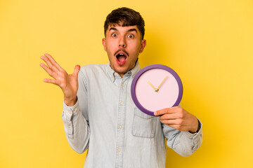 Young hispanic man holding a clock isolated on yellow background surprised and shocked.