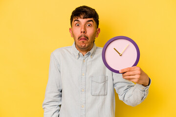Young hispanic man holding a clock isolated on yellow background shrugs shoulders and open eyes confused.
