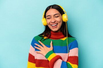 Young hispanic woman listening to music isolated on blue background laughs out loudly keeping hand on chest.