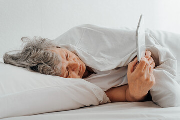 Senior woman covered in blanket using tablet while lying in bed in bedroom. Side view of sleepy mature woman reading online or browsing internet while relaxing in room. Elderly people and technology
