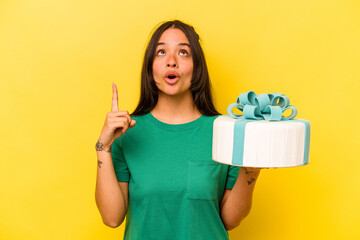 Young hispanic woman holding birthday cake isolated on yellow background pointing upside with opened mouth.