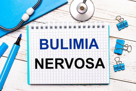 On a light wooden background, a stethoscope, a blue notepad, blue paper clips, a blue marker and a sheet of paper with the text BULIMIA NERVOSA. Medical concept