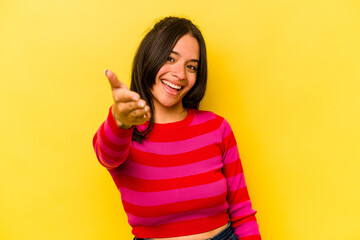 Young hispanic woman isolated on yellow background stretching hand at camera in greeting gesture.