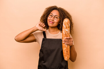 Young baker African woman holding a loaf of bread isolated on beige background feels proud and self...
