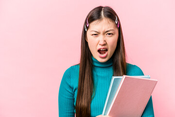 Young asian student woman holding books isolated on pink background screaming very angry and...