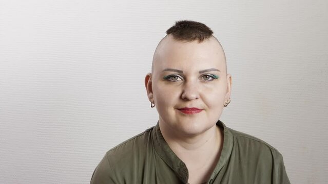 A shaved woman with a mohawk sits on a gray background smiling joyfully. Medium plan