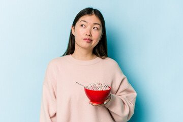 Young asian woman holding a bowl of cereals isolated on blue background dreaming of achieving goals...