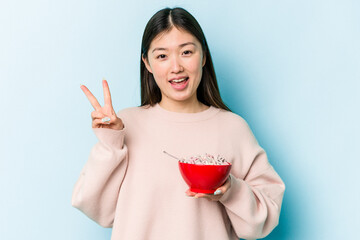 Young asian woman holding a bowl of cereals isolated on blue background joyful and carefree showing a peace symbol with fingers.